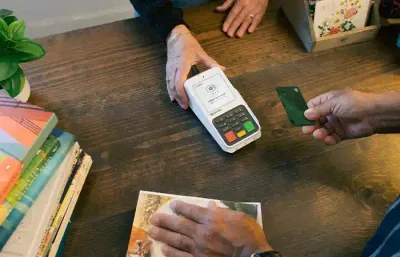 A merchant’s hand pushes a Clover Compact card reader on a corner toward a customer who is extending a credit card to tap pay