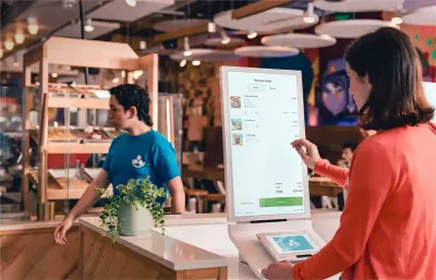 A woman in an orange sweater orders food on a Clover Kiosk point of sale unit in a bakery while a worker passes behind the counter