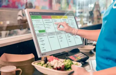 A foodservice worker wearing rubber gloves holds a bowl of salad in one hand while using the other to check orders on a Clover Kitchen Display System screen