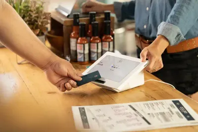 A merchant holds onto a Clover Mini point of sale unit on a wooden counter while a customer’s hand taps a credit card to pay
