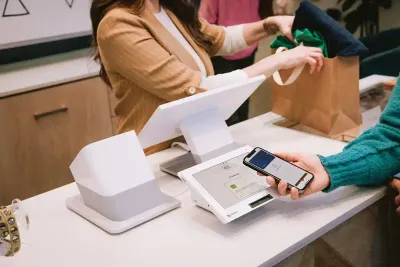 A salesperson places clothing in a shopping bag while a customer taps their phone on a Clover Station Duo point of sale system to pay