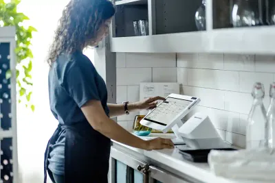A foodservice worker wearing an apron stands in a bright, clean kitchen space using a Clover Station Solo to input a transaction